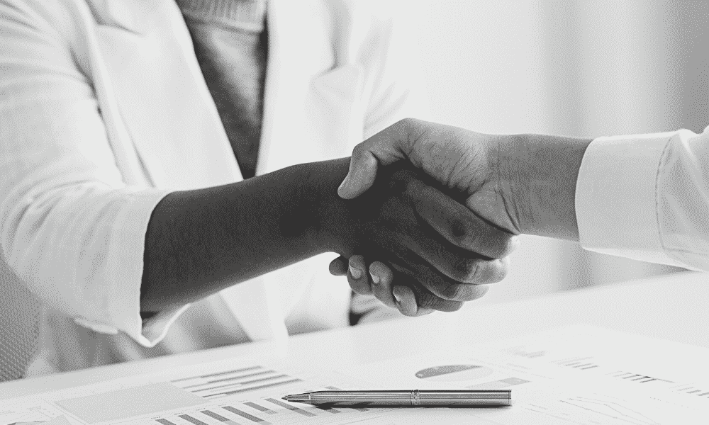 Two business women shake hands over signed paperwork. The woman facing you is black, the woman out of frame is white.
